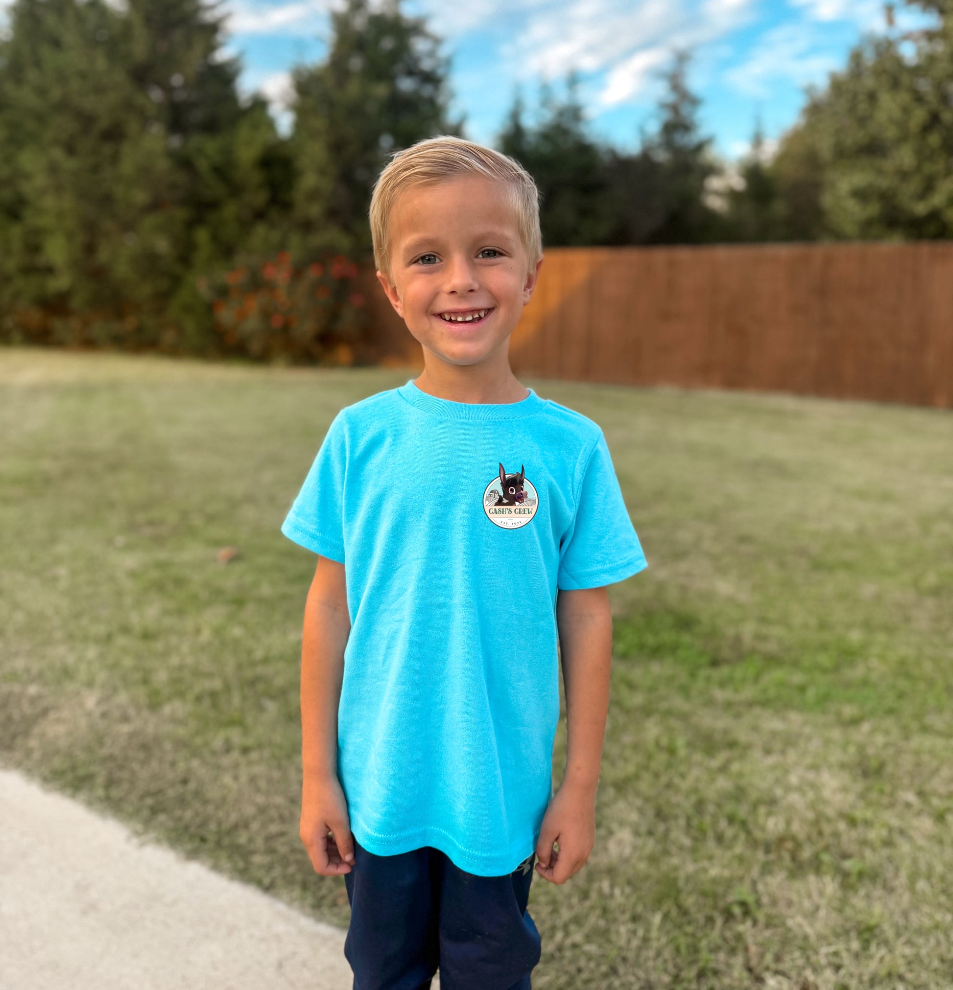 Young boy wearing a blue t-shirt with a logo, standing outdoors on a sunny day.